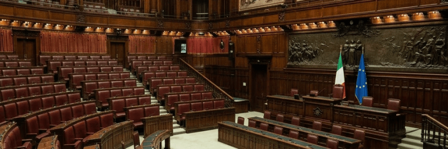 Speaker addressing seated members in the Italian Parliament chamber with Italian and European Union flags and large historic paintings